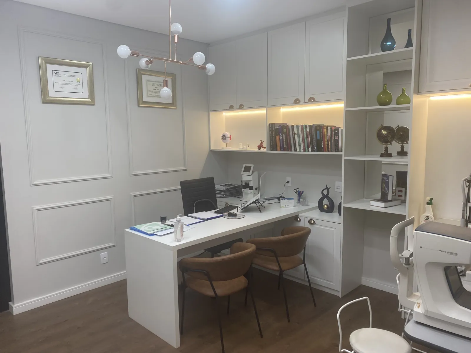 Consultório do Dr. Ruy: mesa branca com computador, estante com livros e diplomas emoldurados na parede
