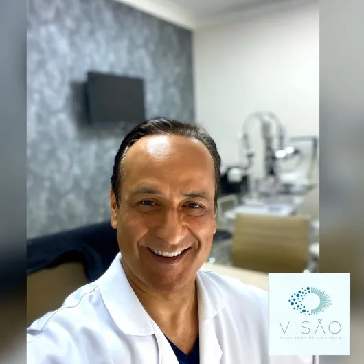 Dr. Ruy dos Santos Filho de jaleco branco, sorrindo, na sala de exames da clínica